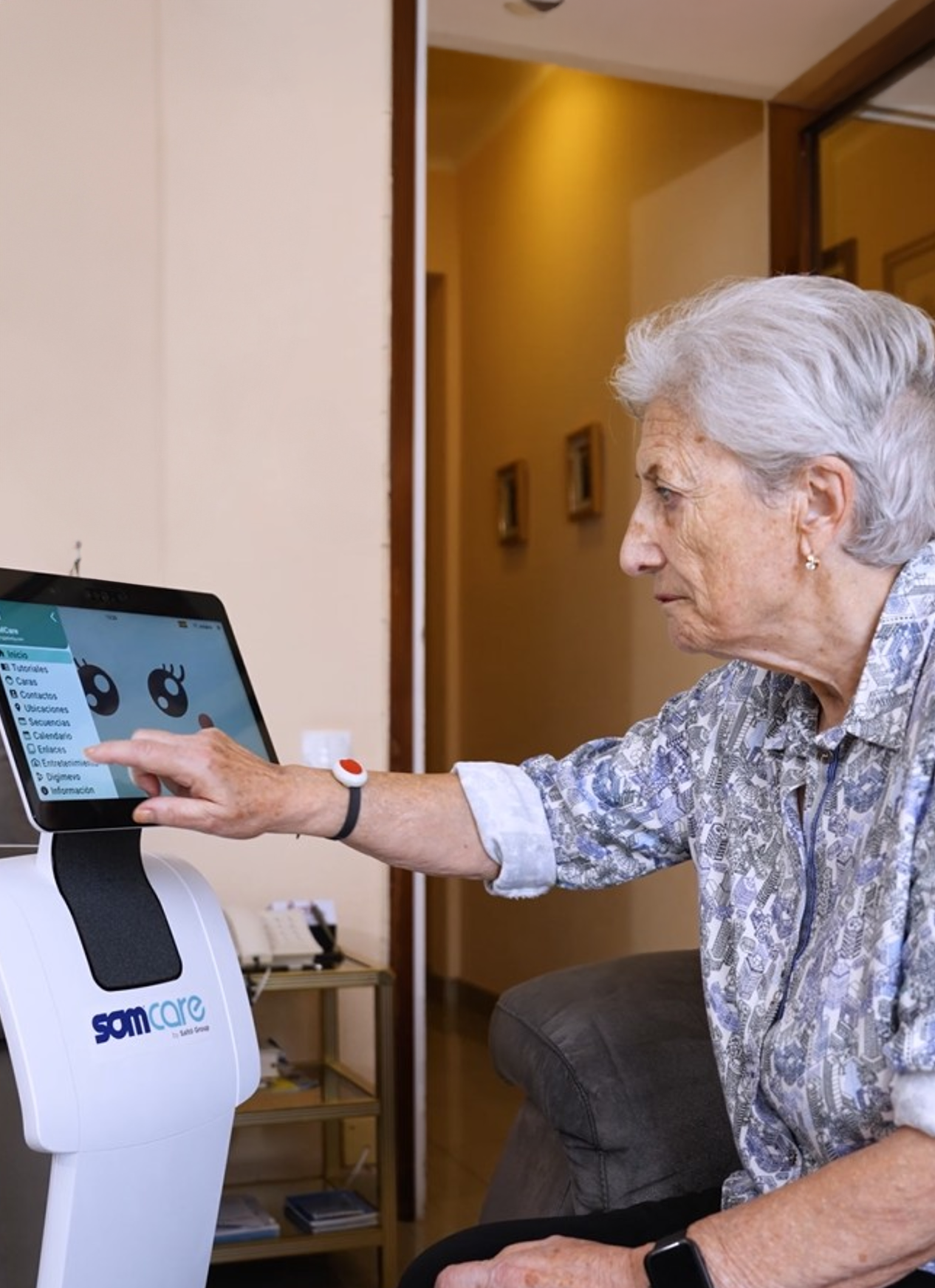 Dona gran interactuant amb un robot assistencial a casa, tocant la pantalla tàctil amb menú d'opcions i avatar animat.