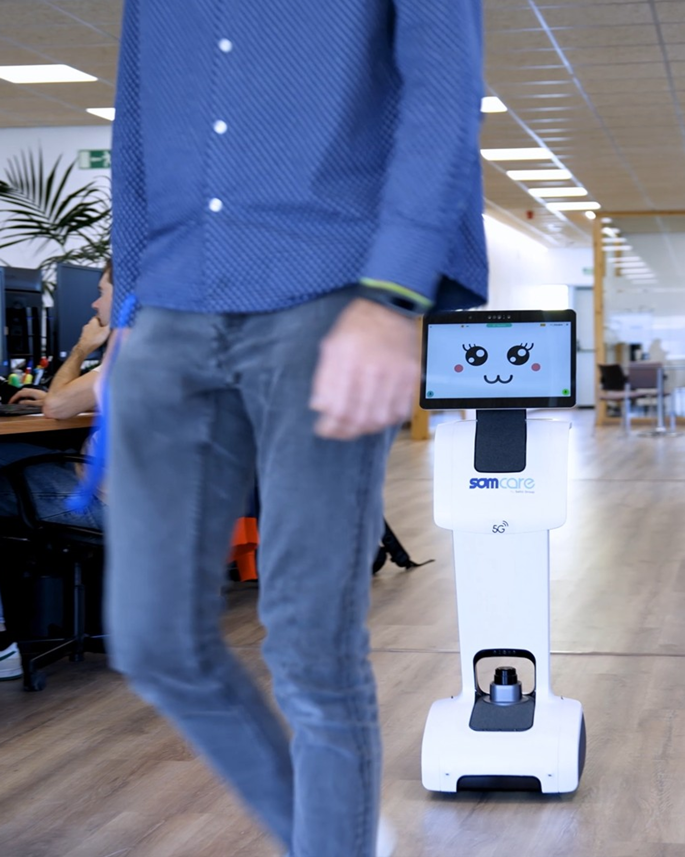 Robot asistencial con pantalla que muestra una cara animada siguiendo a una persona en una oficina de Group Saltó; en el cuerpo del robot se lee “SOMcare” y 5G.