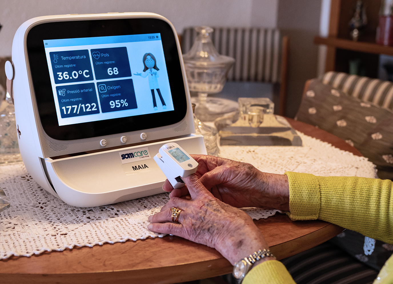 Persona gran utilitzant un dispositiu de telemonitorització a casa amb el dispositiu MAIA; la pantalla mostra temperatura, pols, pressió arterial i nivell d'oxigen.