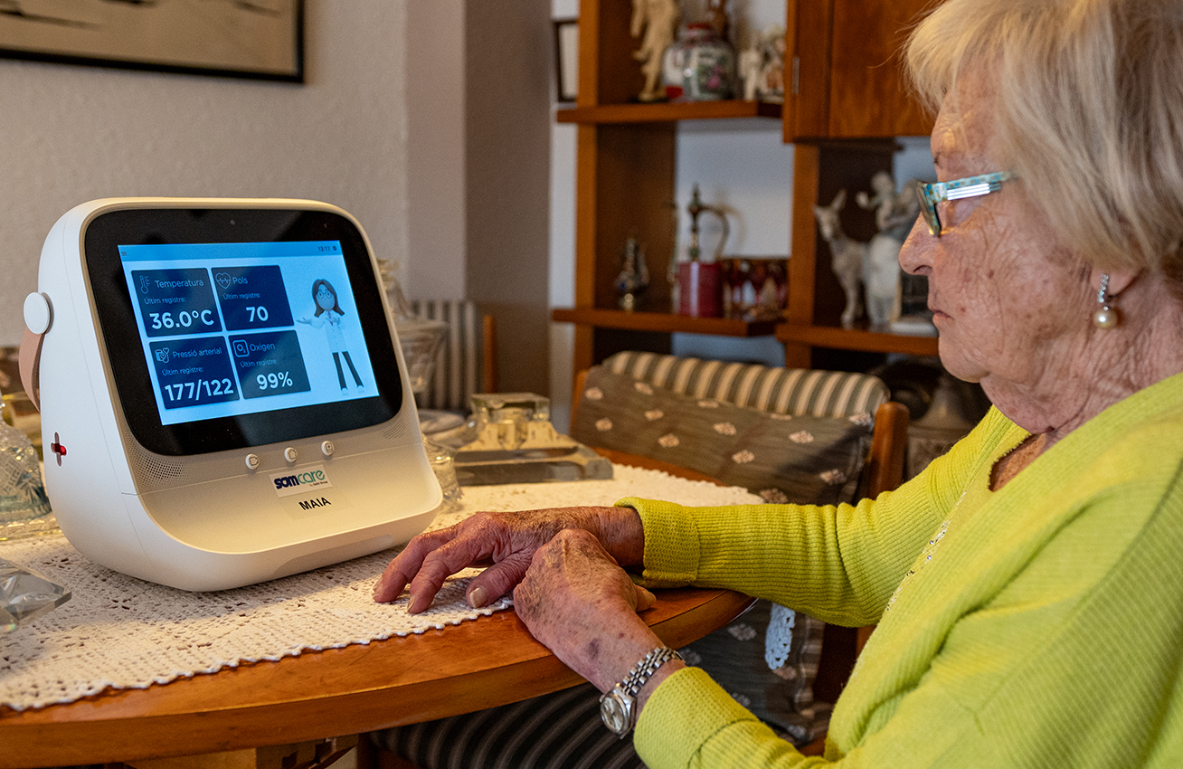 Persona gran utilitzant un dispositiu de telemonitorització a casa; la pantalla mostra temperatura, pols, pressió arterial i nivell d'oxigen.