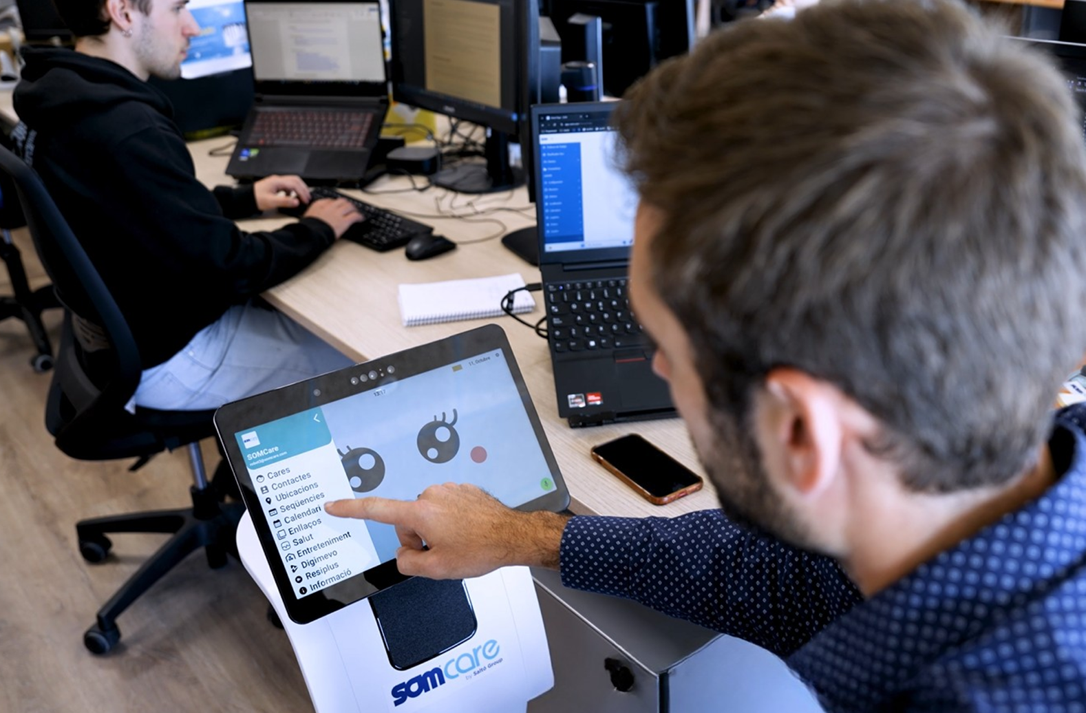 Persona utilitzant la pantalla tàctil dun robot assistencial en una oficina, amb altres treballadors i ordinadors al fons.