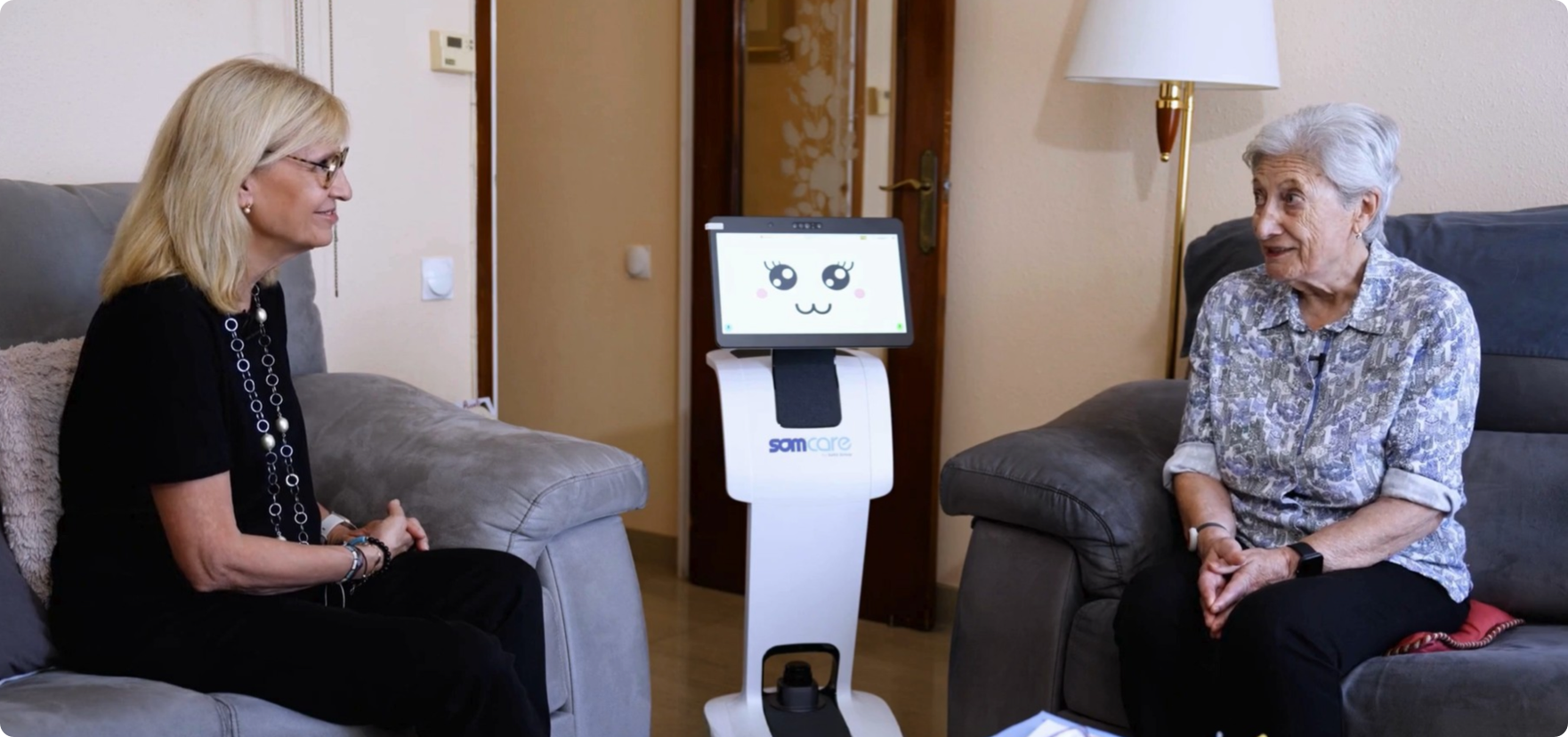 Dues dones assegudes al saló conversant amb un robot assistencial al mig, la pantalla del qual mostra una cara animada.