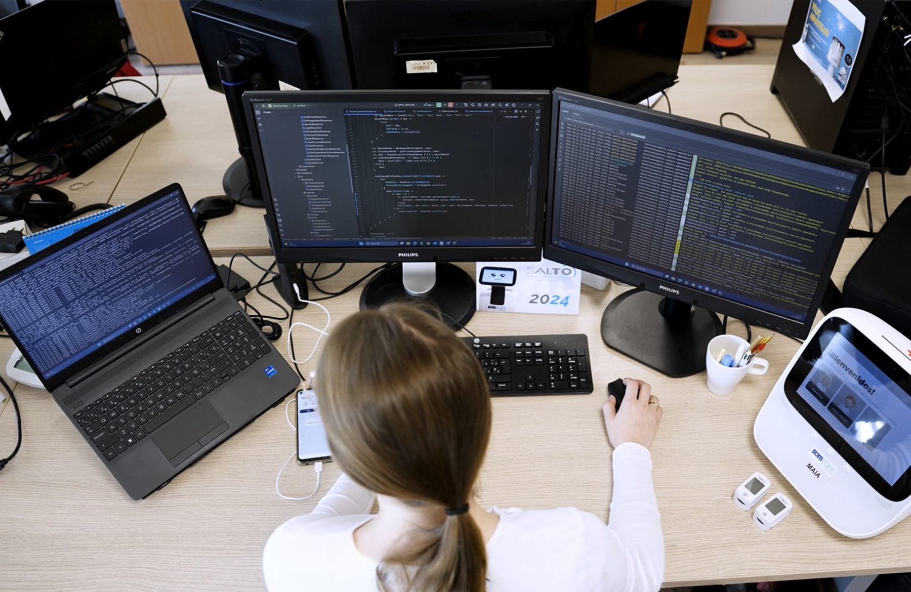 Programadora treballant en un escriptori amb diversos monitors mostrant codi i registres del sistema; de banda hi ha un dispositiu de teleassistència amb pantalla encesa i sensors mèdics.