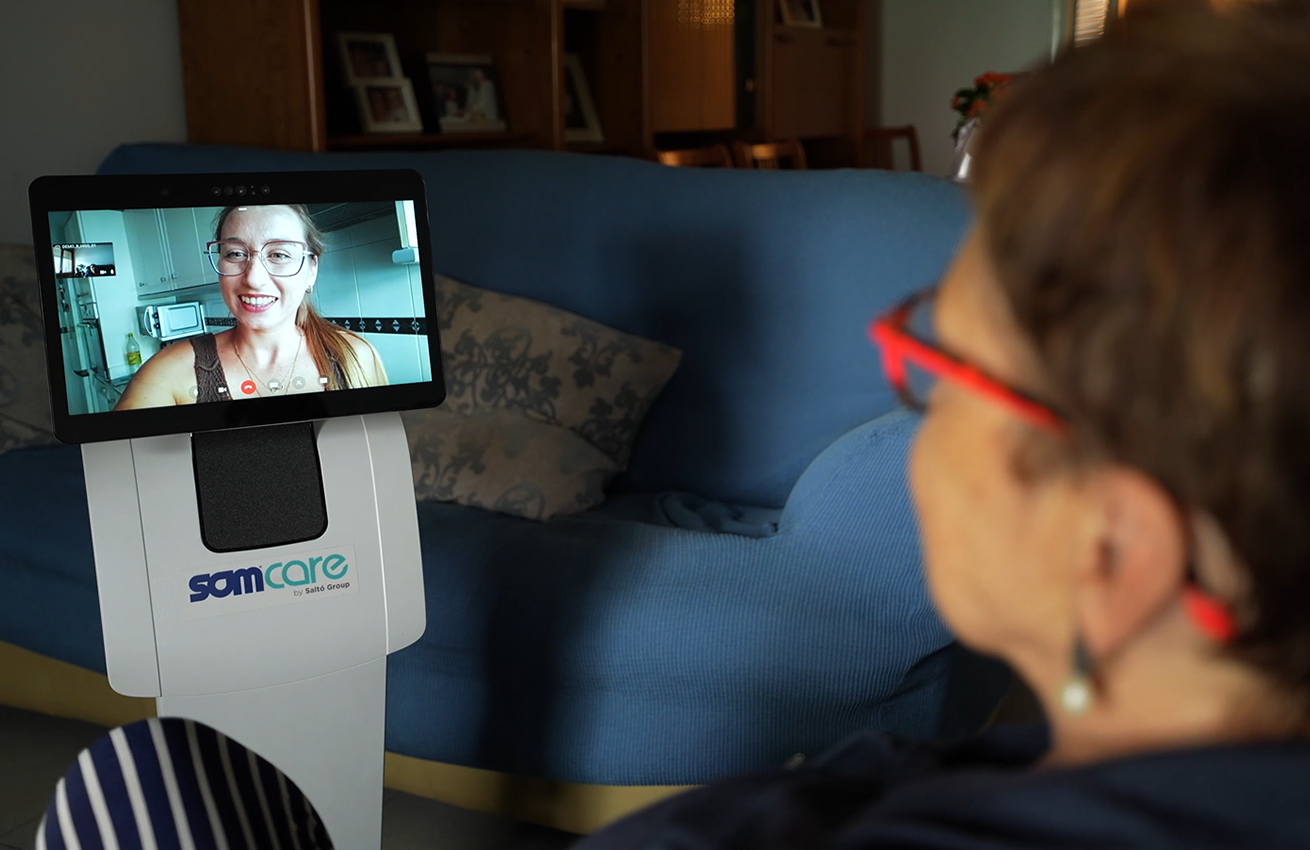 Mujer mayor realizando una videollamada desde casa a través de un robot asistencial con pantalla; en la pantalla aparece una persona sonriendo durante la llamada.